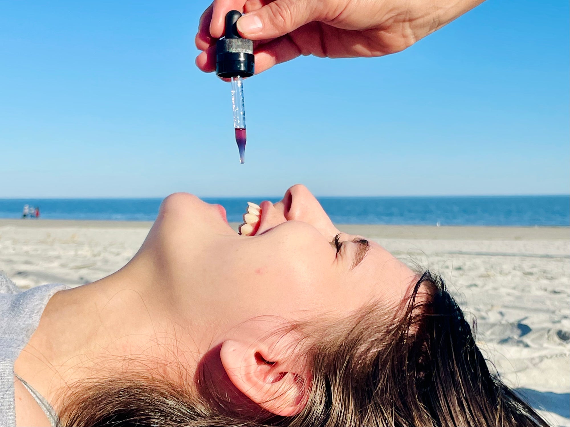 Person lying on a beach while a dropper with liquid is held above their open mouth under a clear blue sky