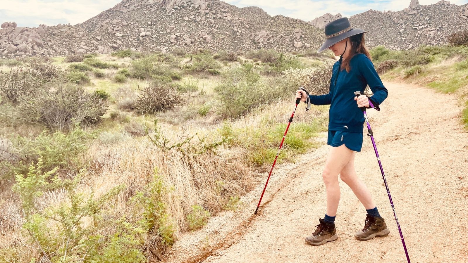 Woman hiking a desert trail with trekking poles, wearing a wide-brim hat and boots among rocky hills and shrubs