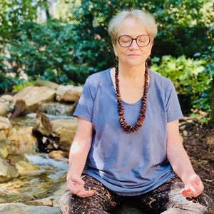 Elderly woman meditating with eyes closed by a stream in a forested area
