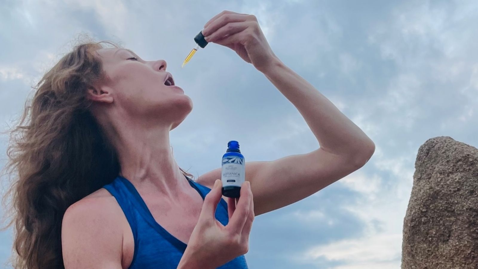 Person outdoors using a dropper to take a liquid herbal supplement, holding a small bottle against a cloudy sky