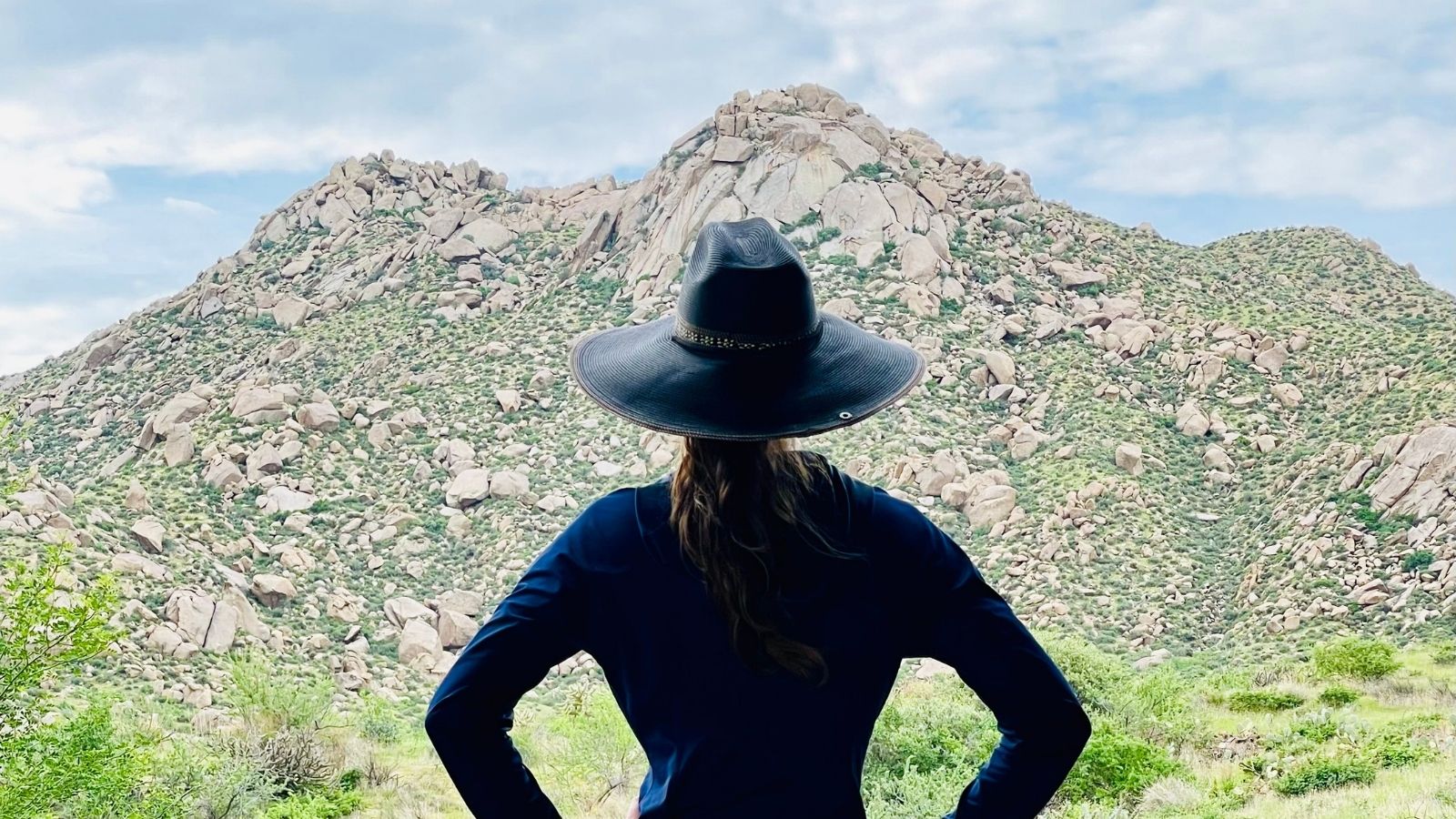 Person wearing a wide-brim hat stands on a desert trail, looking toward rocky green mountains under a cloudy sky