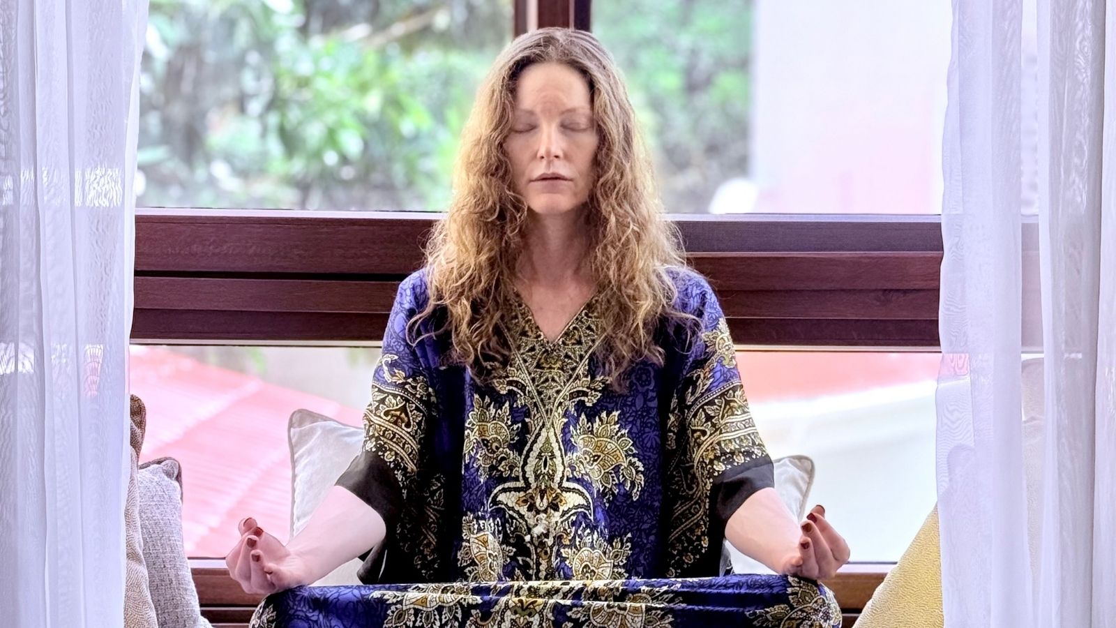 Woman sitting cross-legged indoors with eyes closed, meditating by a window, wearing a patterned robe