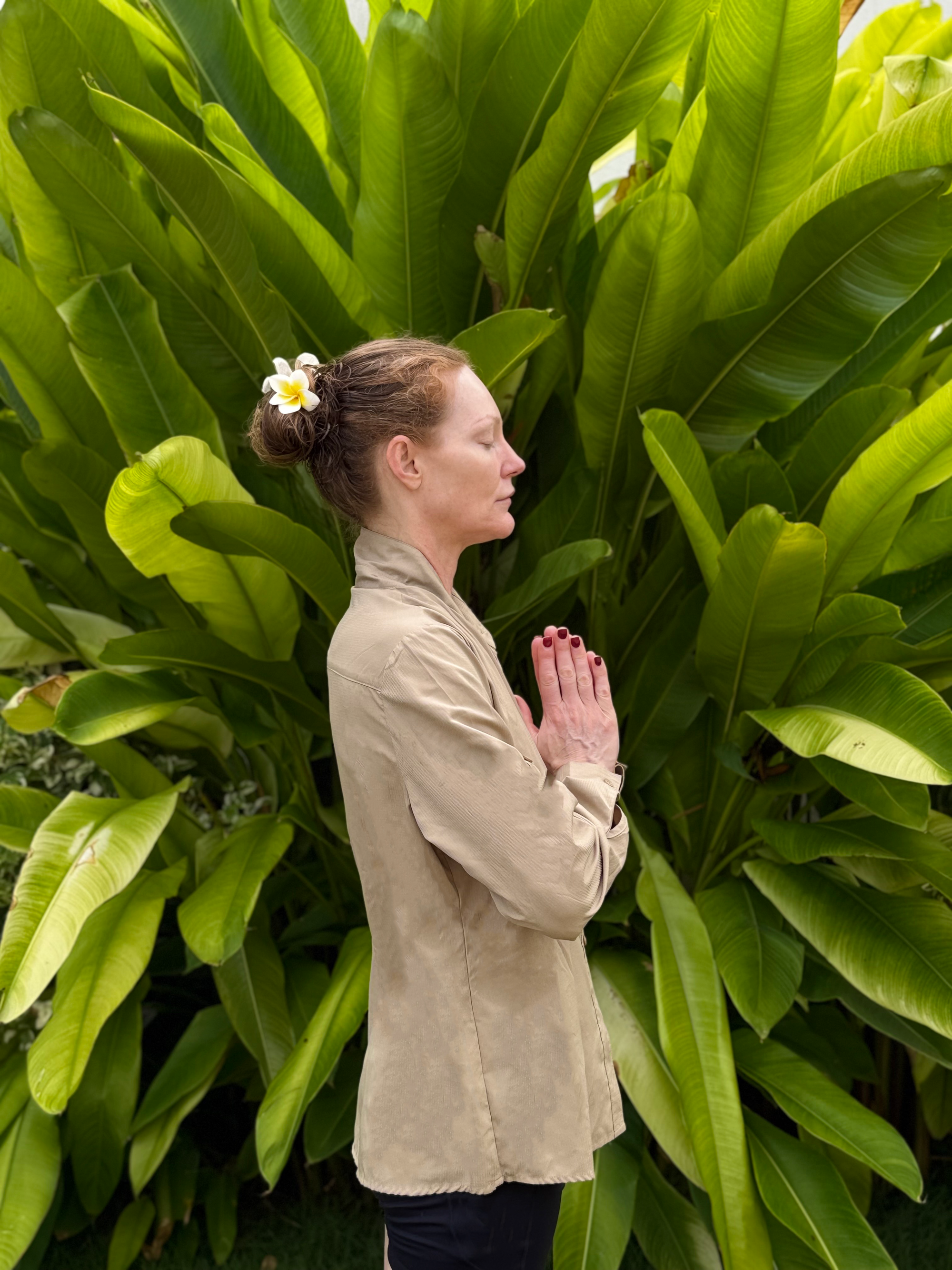 Woman meditating in profile with hands together before lush green tropical leaves
