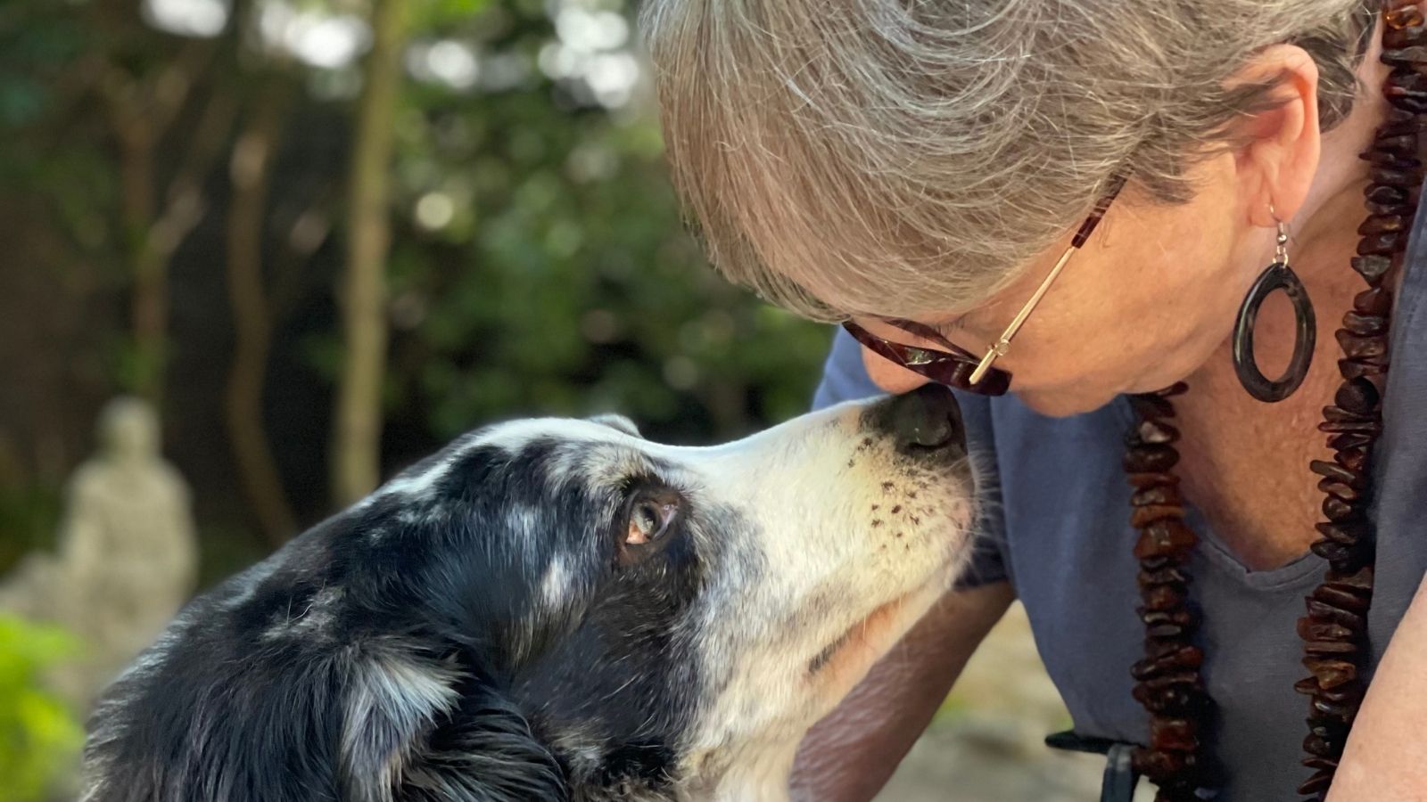 Older woman with glasses gently touching noses with a black-and-white dog outdoors, sharing a quiet, affectionate moment
