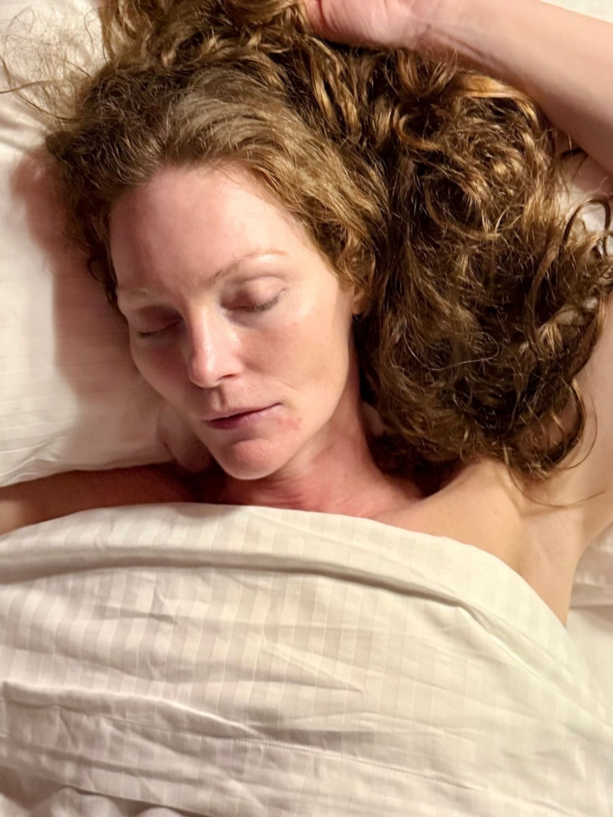 Woman with curly hair resting in bed with eyes closed, lying on white pillows and sheets