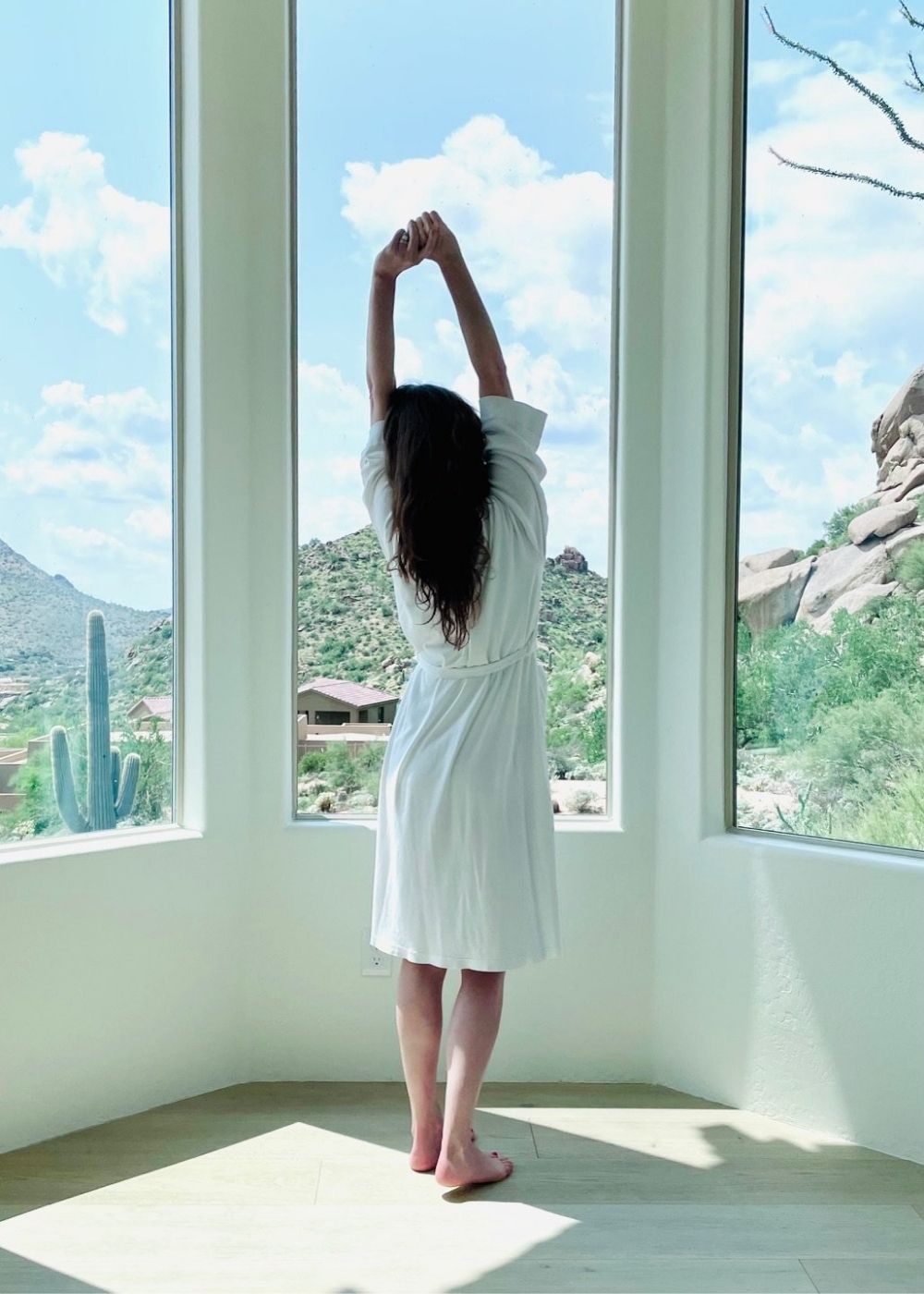 Woman stretching in a white robe by large windows with a desert mountain view.