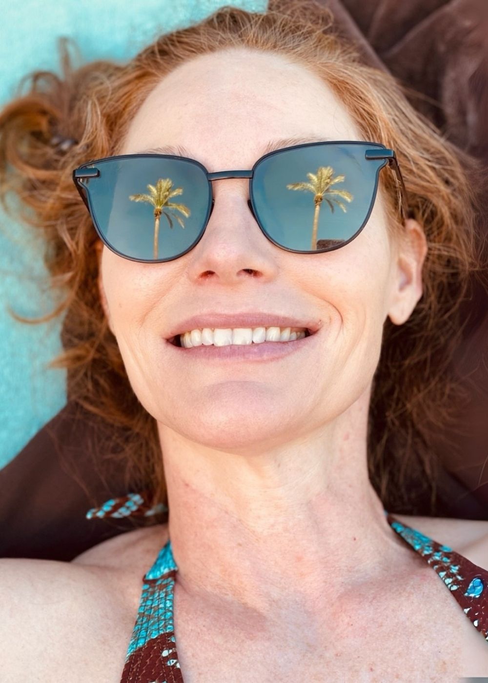 Person smiling while lying down outdoors, with palm trees reflected in their sunglasses