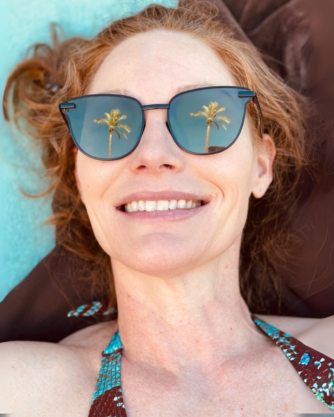 Person smiling while lying down outdoors, with palm trees reflected in their sunglasses