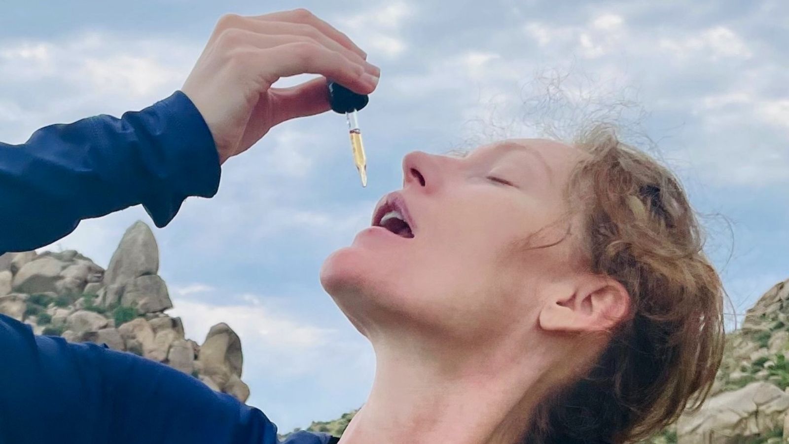 Woman outdoors tilts her head back to take liquid drops from a dropper bottle, with rocky hills and sky behind her